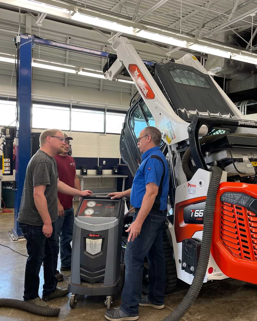 A male instructor and two male students standing in a shop next to a Bobcat